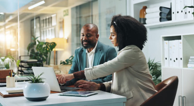 Two people seated at a desk collaborating on a laptop in a modern office setting with plants and shelves in the background