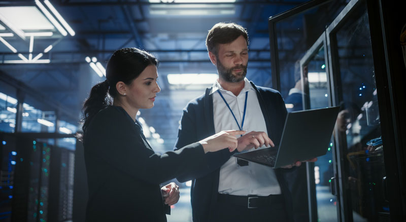 Two IT professionals collaborate in a server room while reviewing data on a laptop.