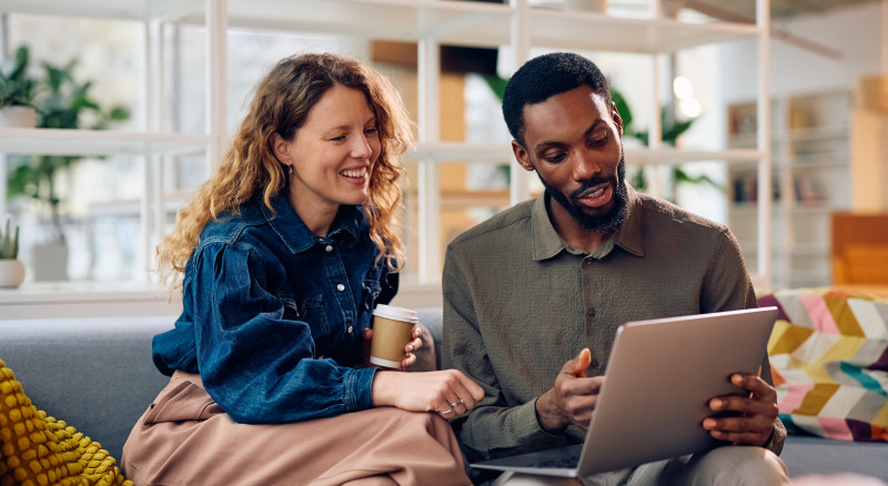 Two people sitting together on a couch looking at a laptop, with one holding a coffee cup.