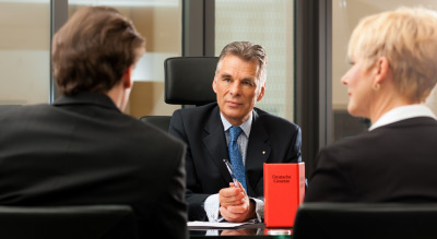 Three people in a meeting, with one holding a pen and a red booklet on the table