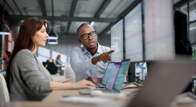 Two coworkers at desks reviewing data on multiple monitors, with one pointing at the screen.
