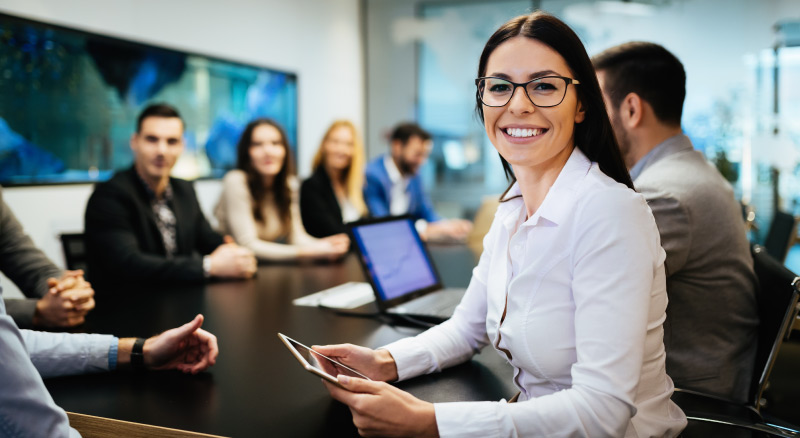 Person at a conference table holding a tablet, with a group meeting in the background
