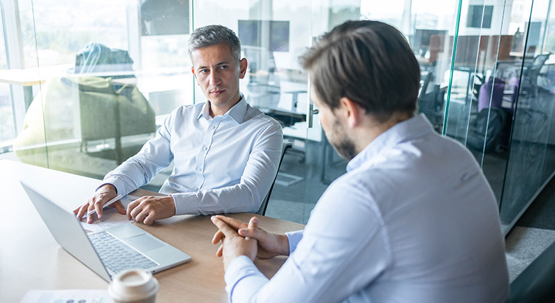 Two colleagues sit at a conference table in a glass-walled office, discussing work while one points at a laptop.