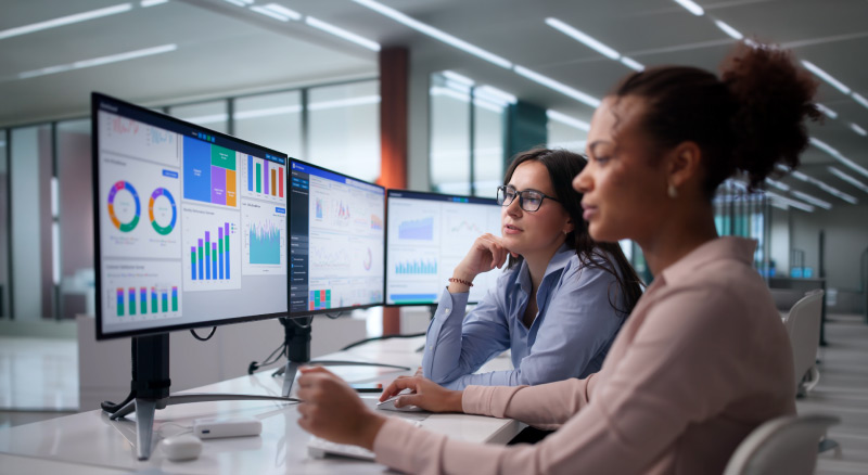 Two people reviewing dashboards on multiple monitors in a modern office.