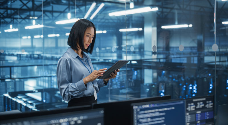 A woman uses a tablet in a modern data center surrounded by computer servers.