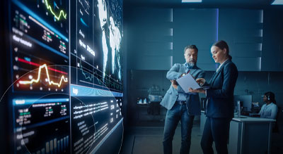 Two professionals review documents in front of large data visualization screens in a technology-focused workspace.