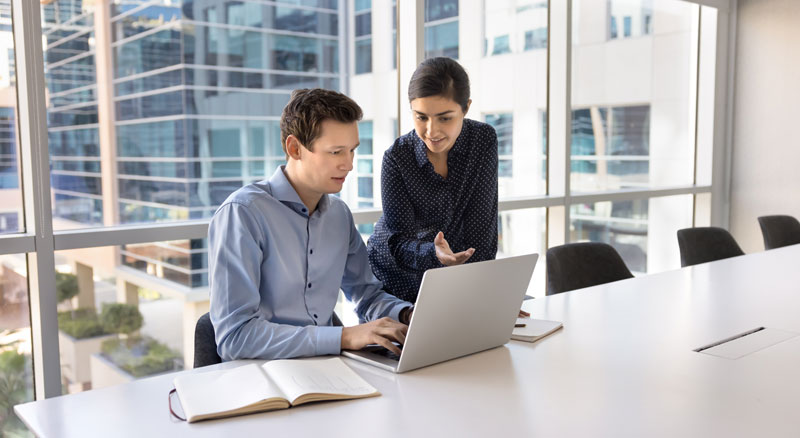 Two colleagues collaborate on a laptop at a conference table in a modern office setting.