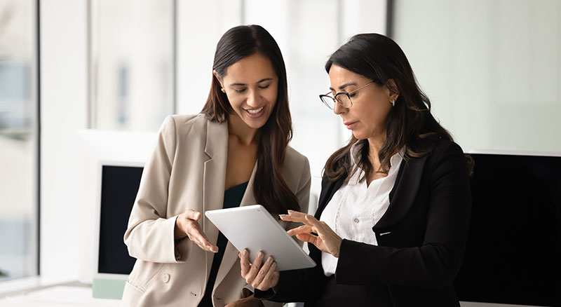 Junior and senior female business colleagues examine content on tablet together.