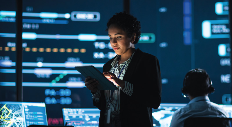 A woman uses a tablet in a dimly lit control room with large digital screens displaying data and graphics in the background.