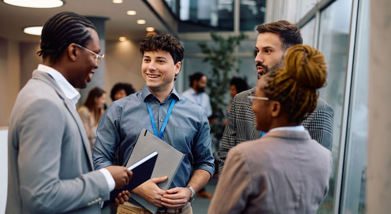 A group of professionals stand in a modern office space, engaged in conversation while holding notebooks and documents