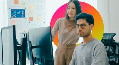 Two coworkers collaborate at a desktop computer in a modern office.