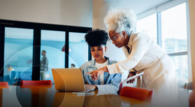 Two people collaborate at a table as one points to a laptop screen in a bright meeting room.