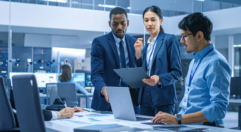 Several professionals gather around a laptop and documents in a technology operations center, collaborating on a project with workstations visible in the background.