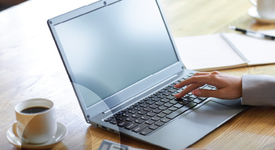A person types on a sleek laptop on a wooden table, with a cup of coffee and an open notebook nearby, creating a productive and serene setting.