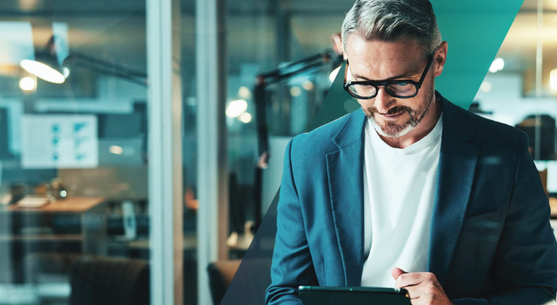 Man with glasses and gray hair wearing a suit reads a tablet in a modern office.