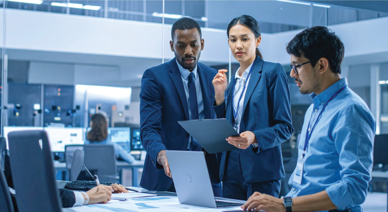 A diverse team of professionals in formal attire collaborates in a modern office, focused on a laptop and documents.