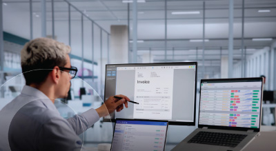 A person in an office setting reviews a digital invoice on a large monitor, with two laptops displaying data.