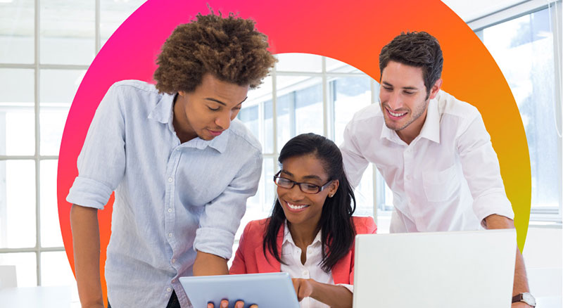Three colleagues collaborate around a laptop and tablet in a modern office setting.