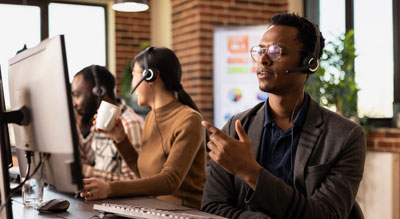 A team of professionals wearing headsets work at computer stations in a modern office environment, collaborating and communicating while using desktop monitors and mobile devices.