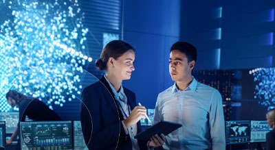 Two professionals collaborate using a tablet in a modern control room with data dashboards and digital displays visible in the background.