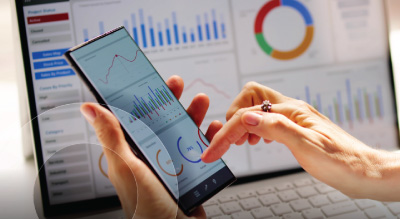 A person interacts with a smartphone displaying graphs while similar charts are visible on a laptop screen in the background, suggesting data analysis.