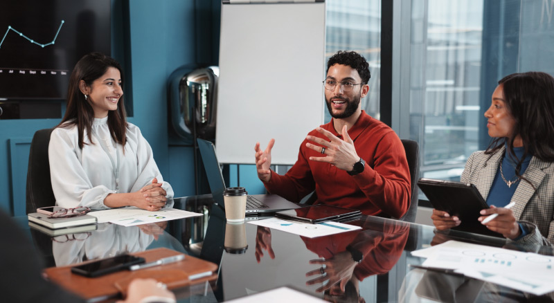 Three coworkers meeting at a table, one speaking while others listen with laptops and documents.