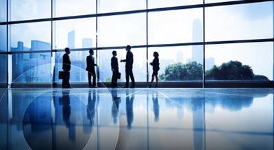Silhouettes of six professionals stand in discussion against a large glass wall with a cityscape view, reflecting a corporate and collaborative atmosphere.