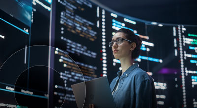 A technology professional stands in front of large digital displays filled with code and data while holding a laptop in a modern operations center.