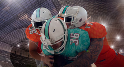 Three football players in teal and orange uniforms collide during a play inside a stadium, with one player being tackled by two defenders.