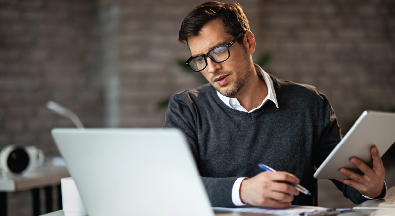Man wearing glasses works at a desk, looking at a laptop screen while holding a tablet and writing with a pen on papers.