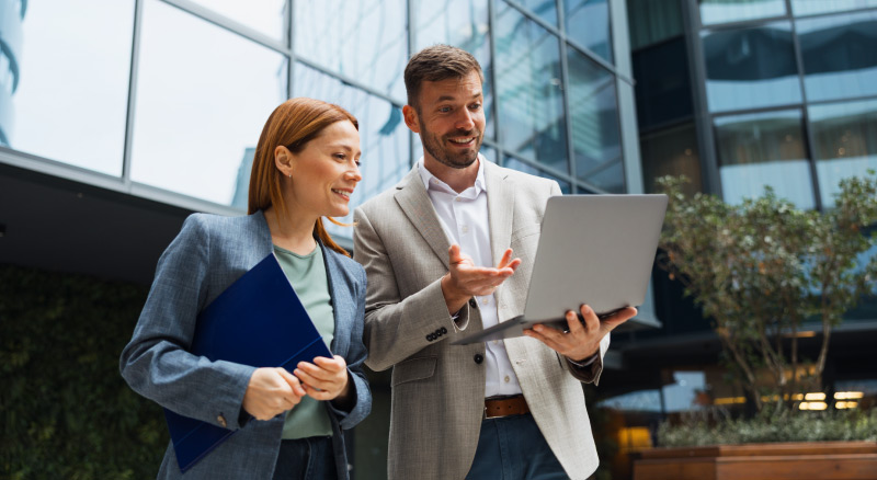 Two people standing outside an office building reviewing a laptop, one holding a folder.
