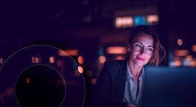 Person working on a laptop in a dimly lit office at night with soft blue and purple lighting.