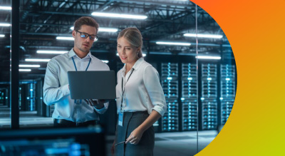 A man and woman in a server room discuss work over a laptop. They appear focused. The background includes server racks. Bright lighting highlights the tech setting.