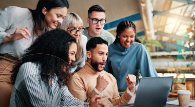 A diverse group of six people excitedly gather around a laptop in an office, looking surprised and delighted.