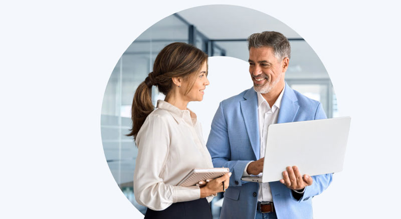 Two colleagues review information on a laptop and tablet while standing in a modern office.