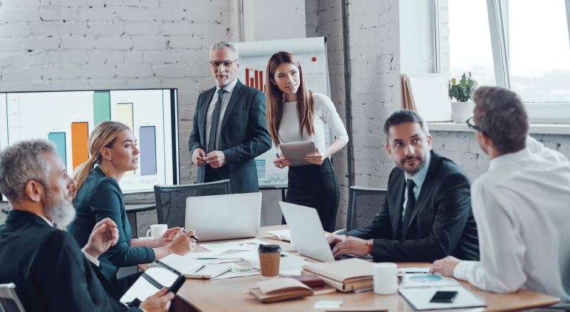 A diverse group of business professionals in a modern office engaged in discussion around a table. A chart and graph are visible in the background.