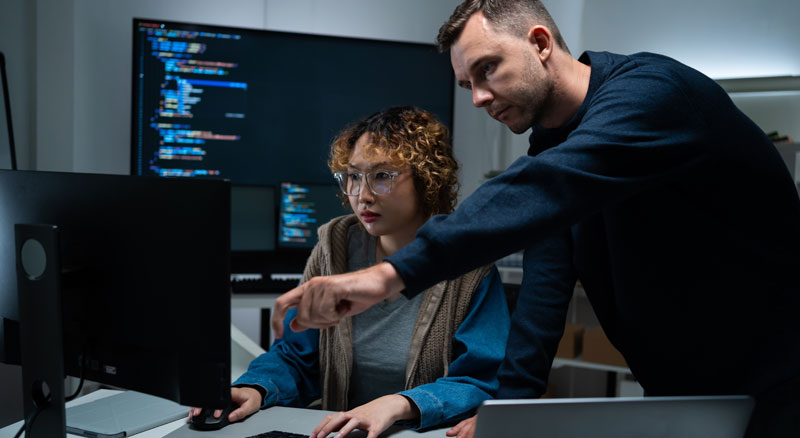 Two software developers focus on code displayed on a monitor in a tech workspace.